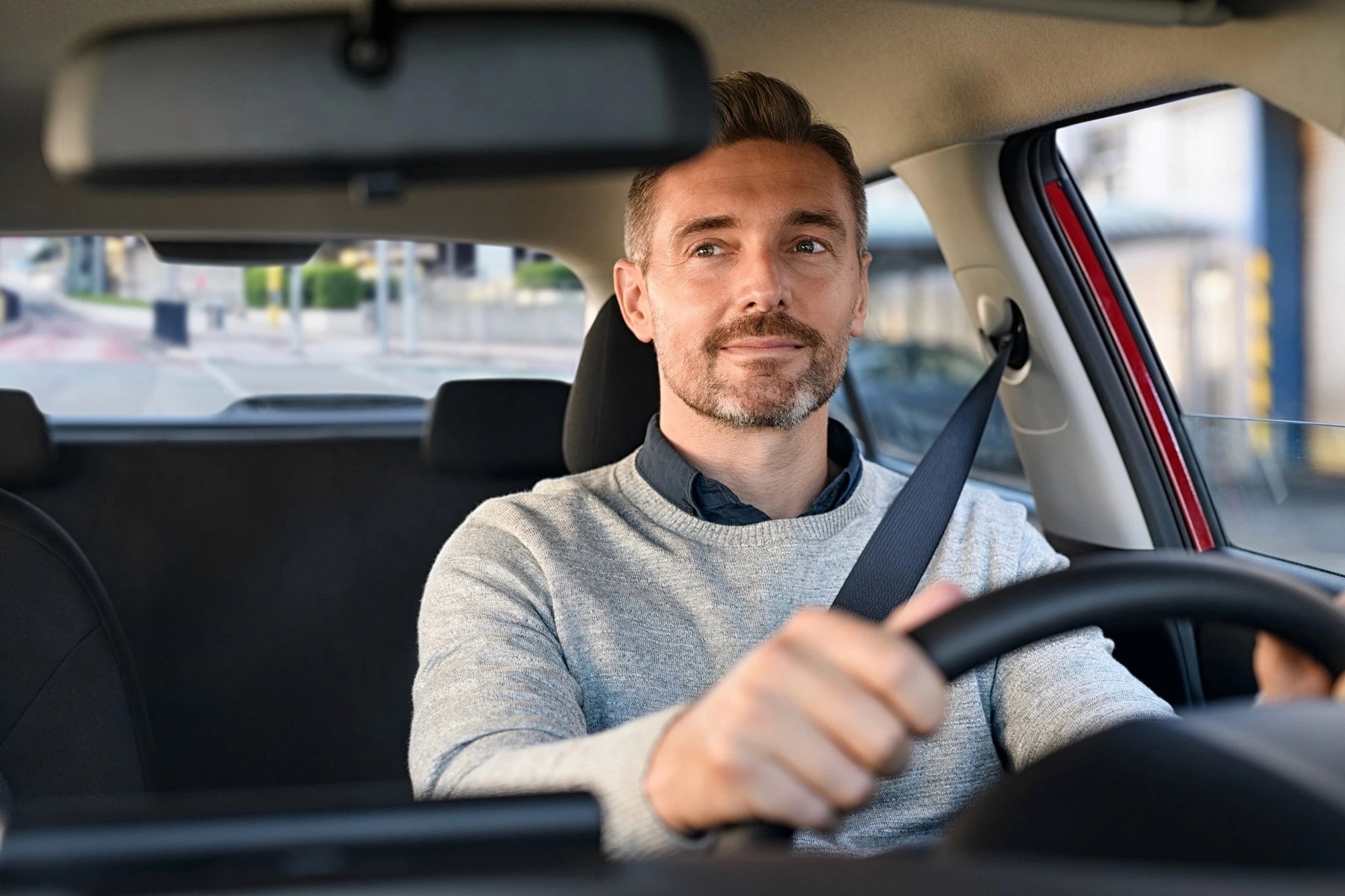 A man driving a car. He is wearing a grey sweater and is looking at the camera with a slight smile.